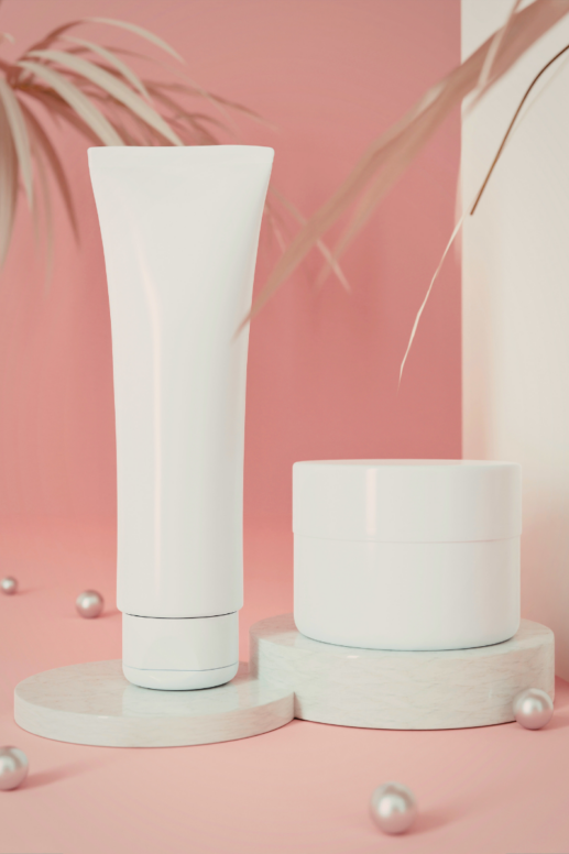 Minimal white skincare tube and jar displayed on marble pedestals against a soft pink background with pearl accents and palm leaves, styled as a clean beauty sunscreen concept image.
