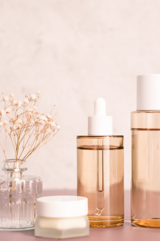 Minimal skincare setup featuring glass bottles with golden-toned serum and dropper, a small cream jar, and dried flowers in a glass vase against a soft neutral background.