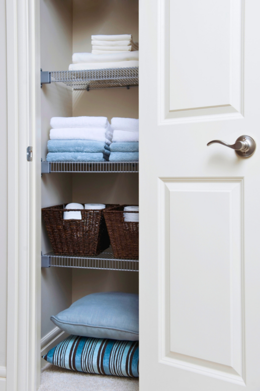 Organized linen closet with neatly folded white and blue towels on wire shelves, woven baskets holding toilet paper, and stacked pillows and blankets, partially visible through an open white door.