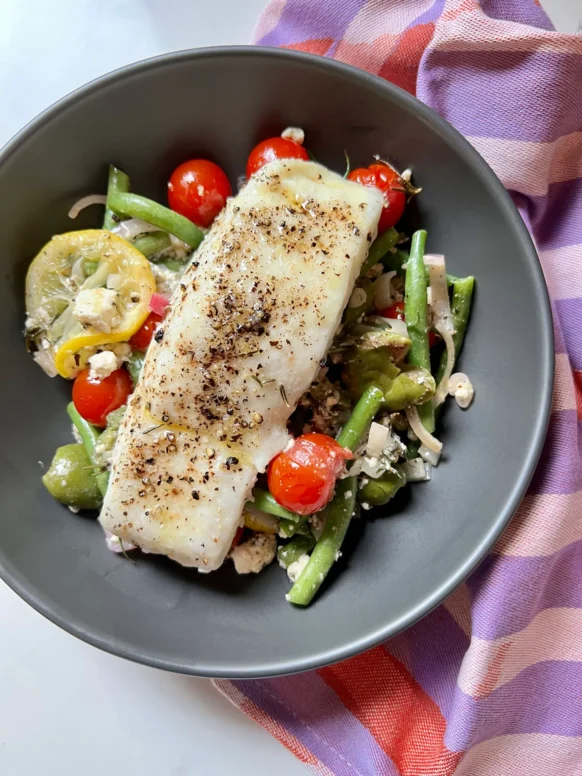Overhead view of plated sheet pan Mediterranean halibut over roasted green beans, tomatoes, olives, feta, and lemon in a dark gray shallow bowl with a colorful checkered napkin