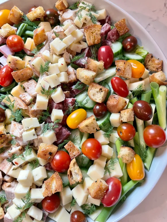 Overhead view of fully assembled chef salad in a wide white bowl with columns of diced meats, cheeses, cucumbers, green beans, scallions, cherry tomatoes, croutons, and fresh dill