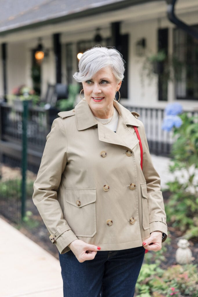 Woman over 50 with silver hair wearing khaki double-breasted cropped trench coat with tortoiseshell buttons over cream sweater, gold hoop earrings, layered gold necklaces, dark wash jeans