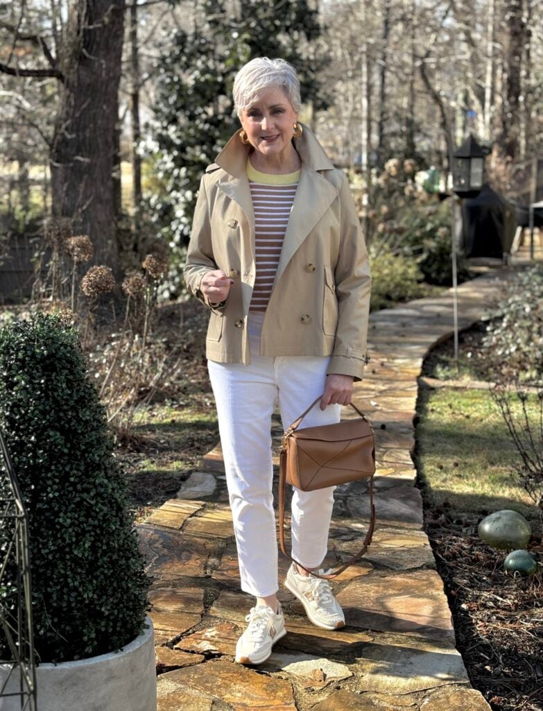 Woman over 50 with silver hair wearing a cropped khaki trench coat over a camel and white striped sweater with green neckline detail, white straight-leg jeans, white leather sneakers, cognac patchwork leather crossbody bag, and gold hoop earrings, standing on a stone garden pathway in early spring