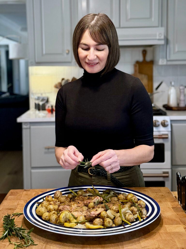 Kelly stands at a butcher block island in a bright kitchen, garnishing a large blue-striped serving platter of roasted lemon herb chicken thighs, baby potatoes, asparagus, and sweet onions with fresh rosemary sprigs.