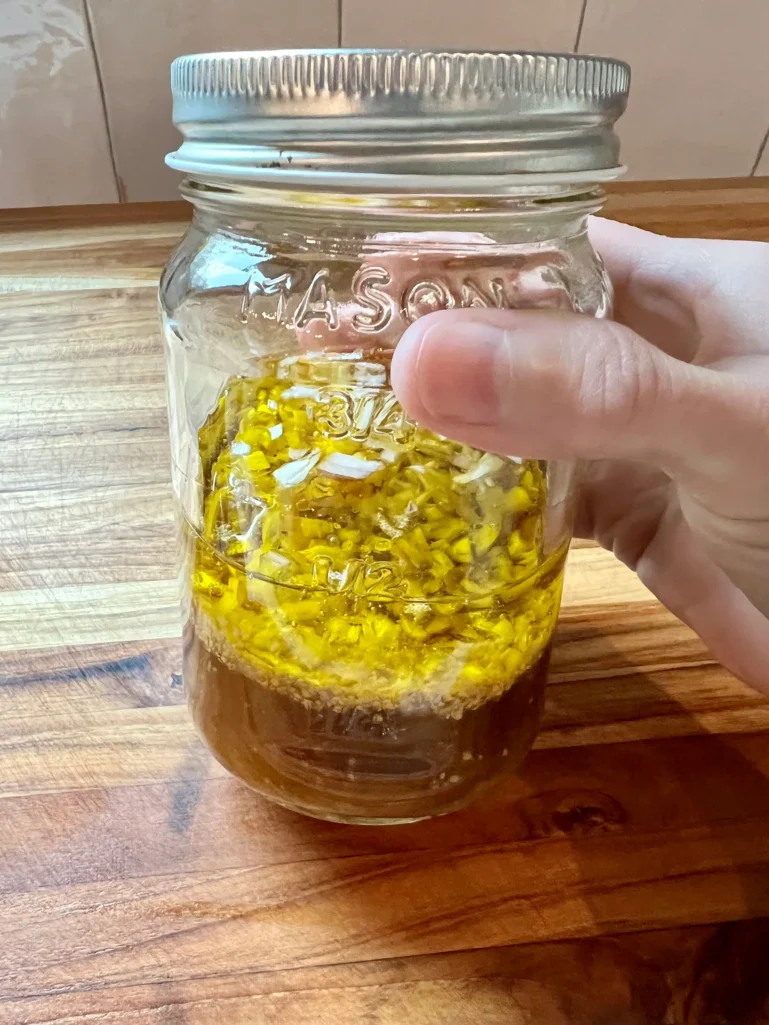 Hand holding a mason jar of shallot vinaigrette with olive oil, white balsamic vinegar, and diced shallots on a wooden cutting board