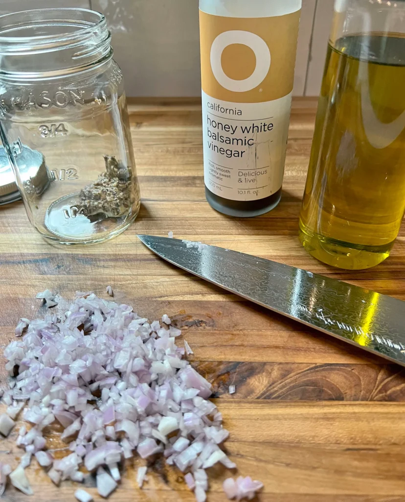 Diced shallot on a wooden cutting board with a mason jar, honey white balsamic vinegar, olive oil, and a chef's knife