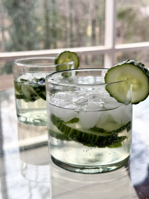 Two Cucumber Gin & Tonics in rocks glasses with cucumber wheels and fresh mint, bubbles catching the afternoon light on the porch table