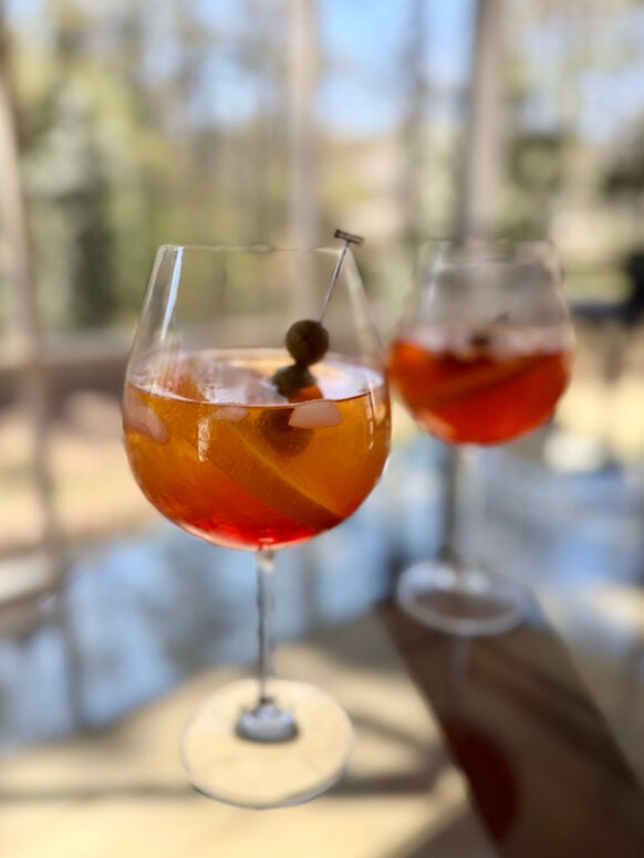 Two Aperol Spritz cocktails in large stemmed wine glasses sit on a sun-drenched table on a screened porch, each garnished with an orange slice and a green olive on a cocktail pick.