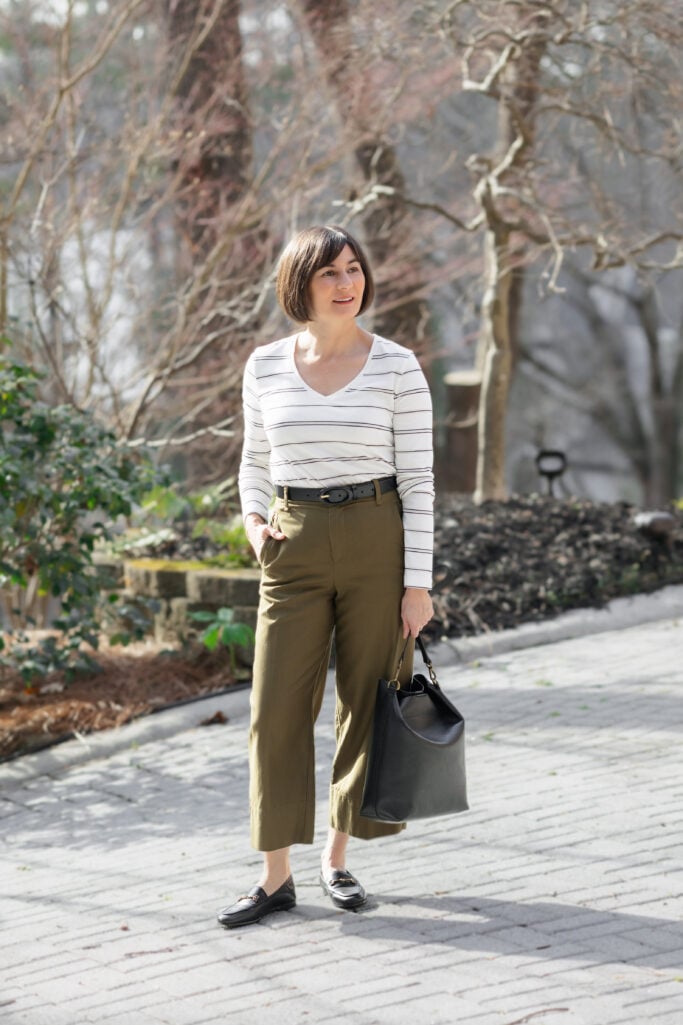 Kelly standing with hand in pocket wearing striped v-neck tee tucked into green wide-leg cropped twill pants with black belt and Loraine loafers — full-length view showing balanced proportions for petite wide-leg styling