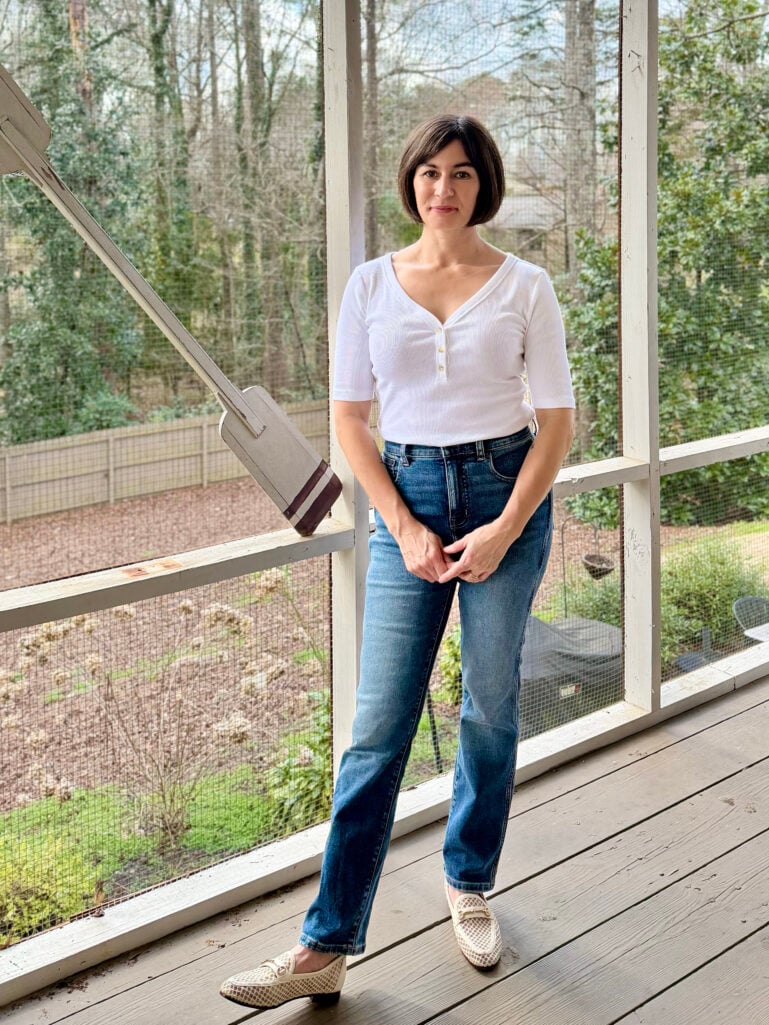 Kelly wearing a white Ribbed Elbow Sleeve Henley Tee, Madewell Petite Pencil Jeans in a medium wash, and Loraine Mesh Bit Loafers, hands clasped on the screened porch