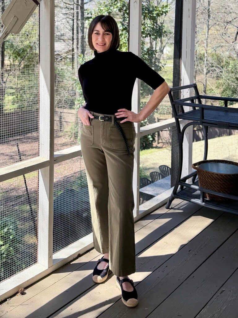 Kelly stands on the screened porch in a black short-sleeve turtleneck, olive wide-leg trousers with a braided leather belt, and black espadrille Mary Janes, one hand on her hip.