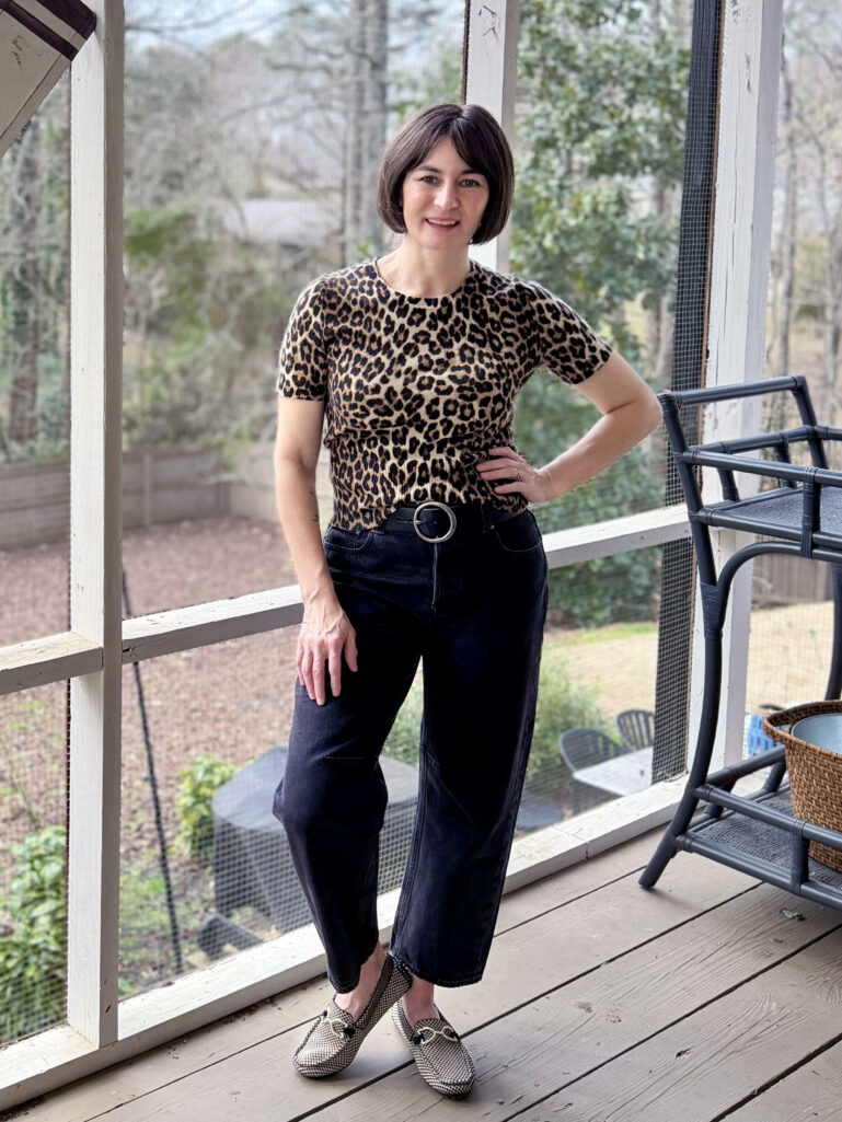 Kelly wearing a leopard print short sleeve sweater, dark darted barrel leg jeans, a round buckle wide belt, and houndstooth Rothy's Daily Driver Loafers on the screened porch, hand on hip