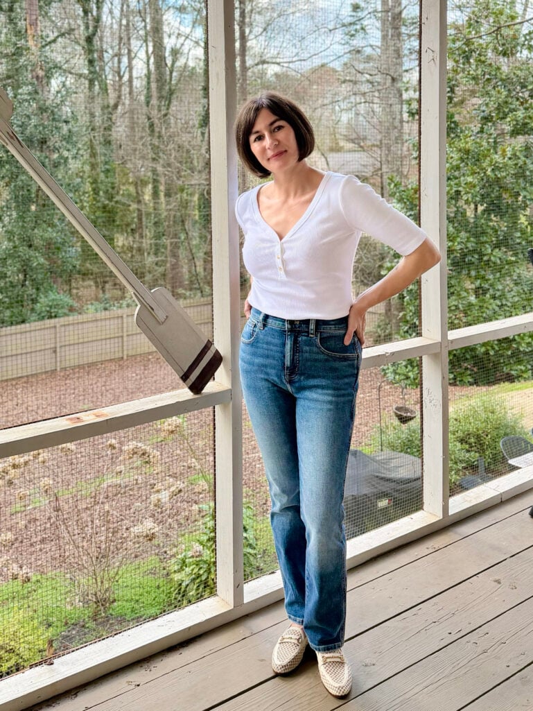 Full-length view of Kelly in her white henley and pencil jeans, hand on hip, leaning against the porch railing in the spring sunshine