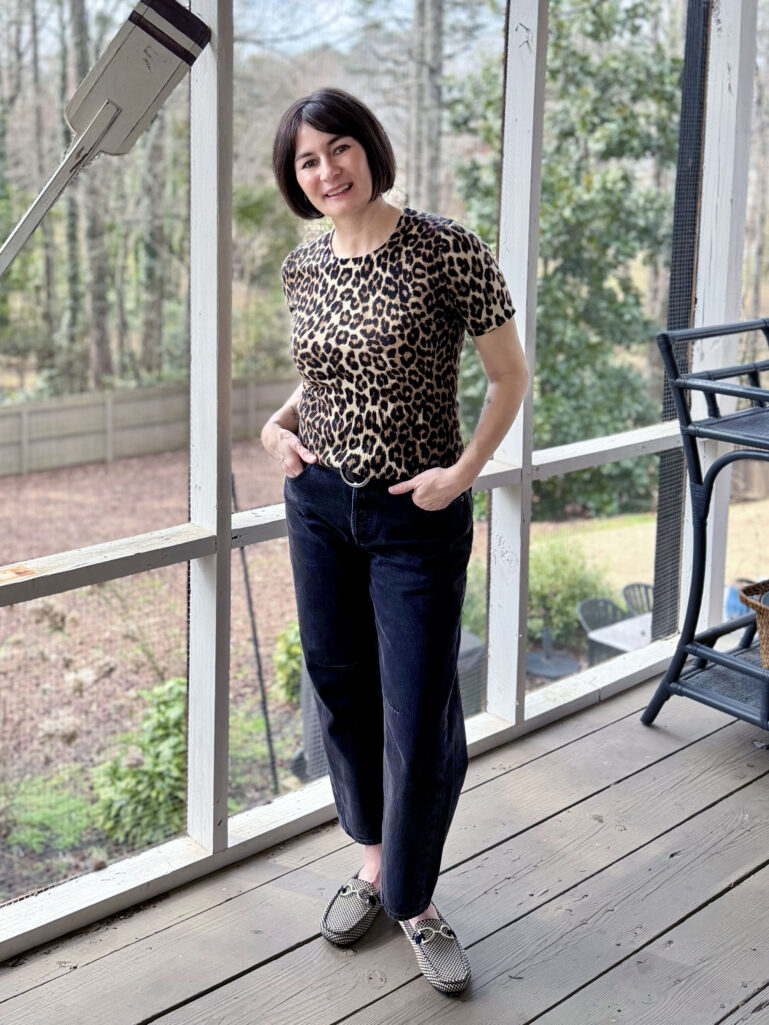 Full-length view of Kelly in her leopard sweater and dark barrel jeans, thumbs in pockets, smiling in the spring light on the porch