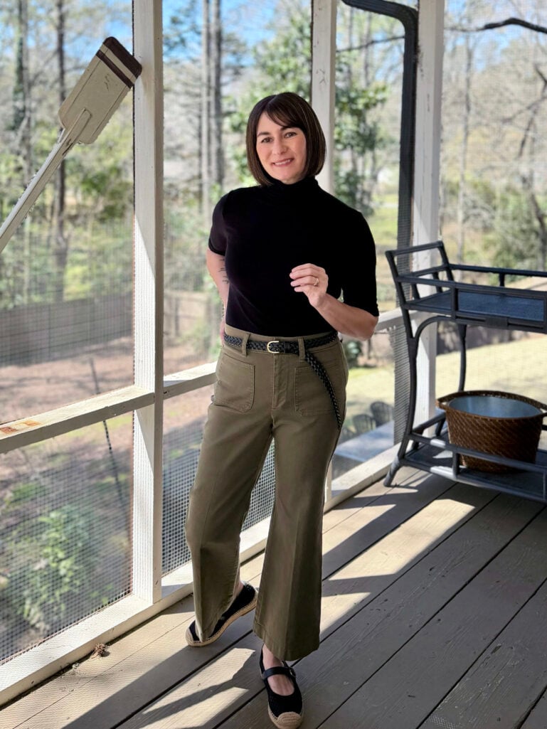 Kelly smiles at the camera on the screened porch, wearing a black short-sleeve turtleneck, olive wide-leg trousers with a braided black belt, and black espadrille Mary Janes.