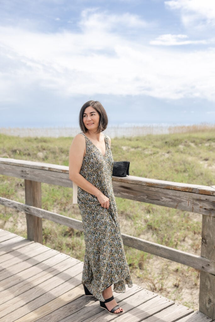 Kelly in Banana Republic Pintuck Maxi Dress on Tybee Island boardwalk, full-length shot showing earth-tone abstract print and black heeled sandals, sea oats and cloudy sky in background — petite spring maxi dress