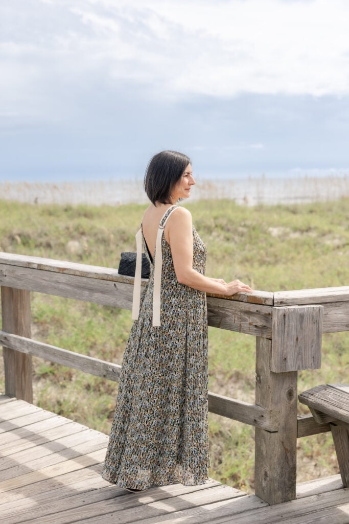 Back view of Kelly in Banana Republic Pintuck Maxi Dress showing cream ribbon strap tie detail and flowing abstract print skirt, standing on Tybee Island boardwalk gazing toward the ocean