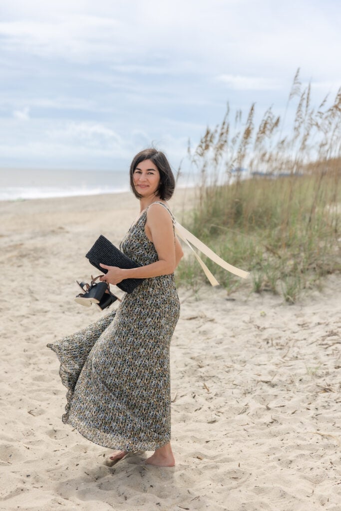 Kelly walking barefoot on Tybee Island beach in flowing Banana Republic Pintuck Maxi Dress, cream ribbon ties streaming in the ocean breeze, holding shoes and black clutch — petite beach dress
