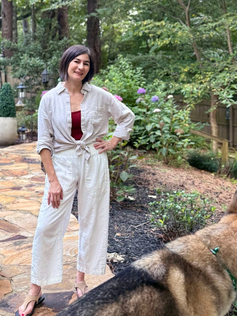 Woman with brown bob haircut wearing oatmeal linen button-down shirt tied at waist over burgundy tank with cream linen pull-on pants, tan wedge sandals