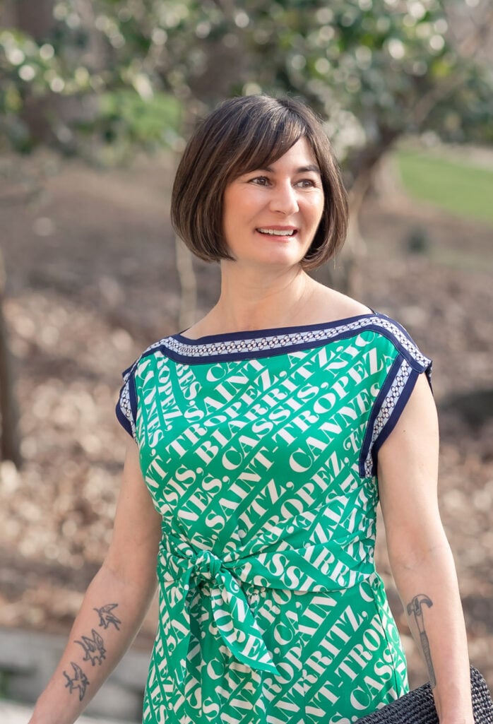 Portrait of Kelly in Anthropologie Maeve Midi Dress in green city-name print, smiling outdoors with bokeh background, showing navy trim neckline and shoulder detail — petite spring dress