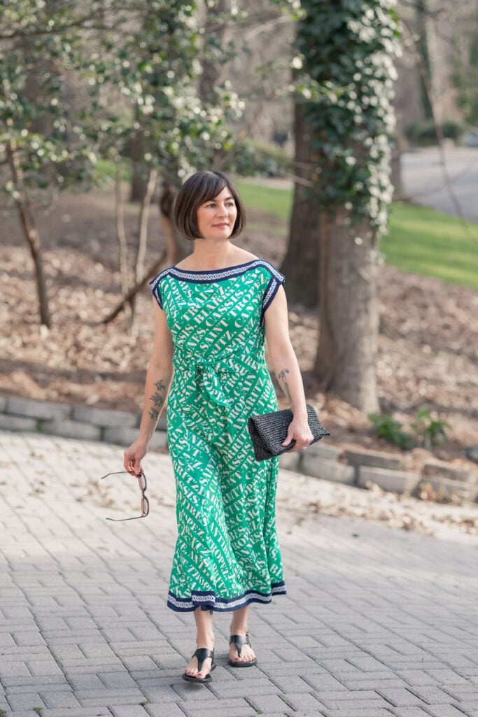 Full-length shot of Kelly in Anthropologie green Maeve Hanky Hem Midi Dress on stone path, showing waist tie detail, navy border hem, and black covered thong sandals, holding sunglasses