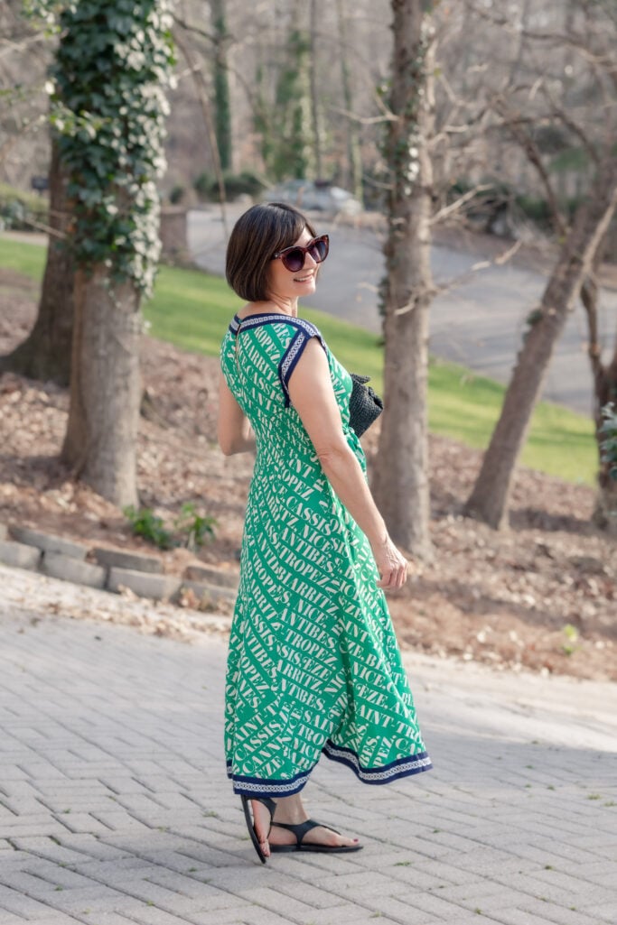 Back view of Kelly in Anthropologie Maeve Hanky Hem Midi Dress showing navy shoulder trim and back silhouette, holding black woven clutch, spring outdoor setting — petite maxi dress back view