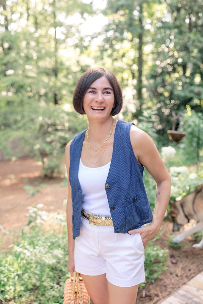 Woman with brown bob wearing navy blue denim vest over white tank top with white shorts, layered gold necklaces, woven belt with gold buckle, woven straw bag