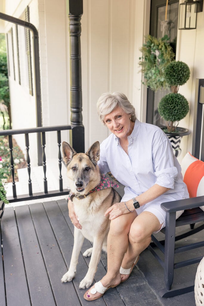 Beth sits in a black rocking chair on the front porch, smiling and holding Oscar, a large German shepherd wearing a bandana and collar, who sits beside her. Topiary ball trees and a lantern are visible in the background.