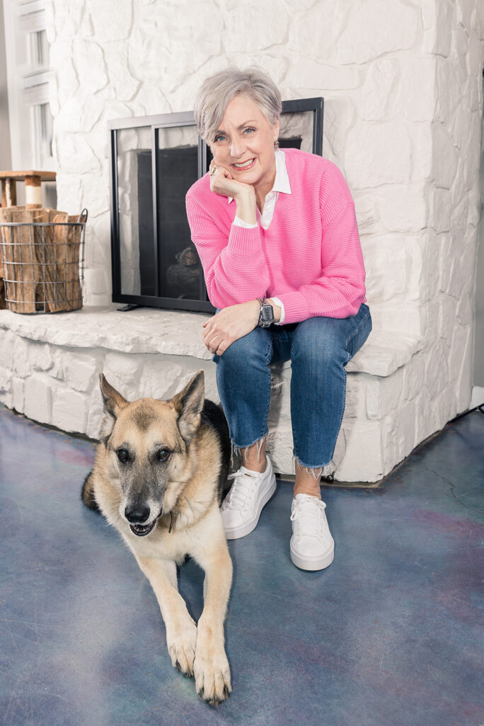 Beth sitting on the stone fireplace hearth in a pink sweater with Oscar the German Shepherd lying at her feet, both looking at the camera, a cozy fireside moment from the archives