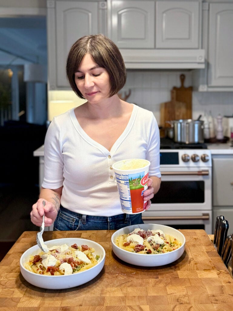 Kelly dolloping Galbani ricotta from the container onto two bowls of Spring Pea Pasta with crispy prosciutto in the kitchen, wearing her white henley tee
