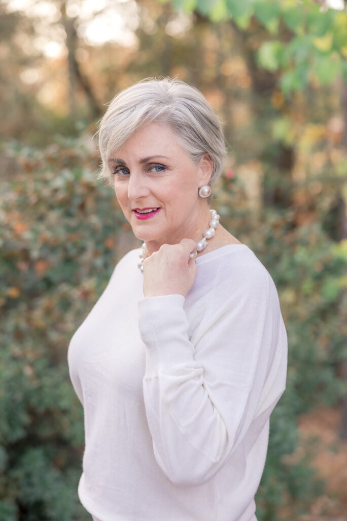 Close-up of woman over 50 with silver hair wearing ivory dolman-sleeve sweater with large pearl drop earrings and chunky pearl bracelet, pink lipstick
