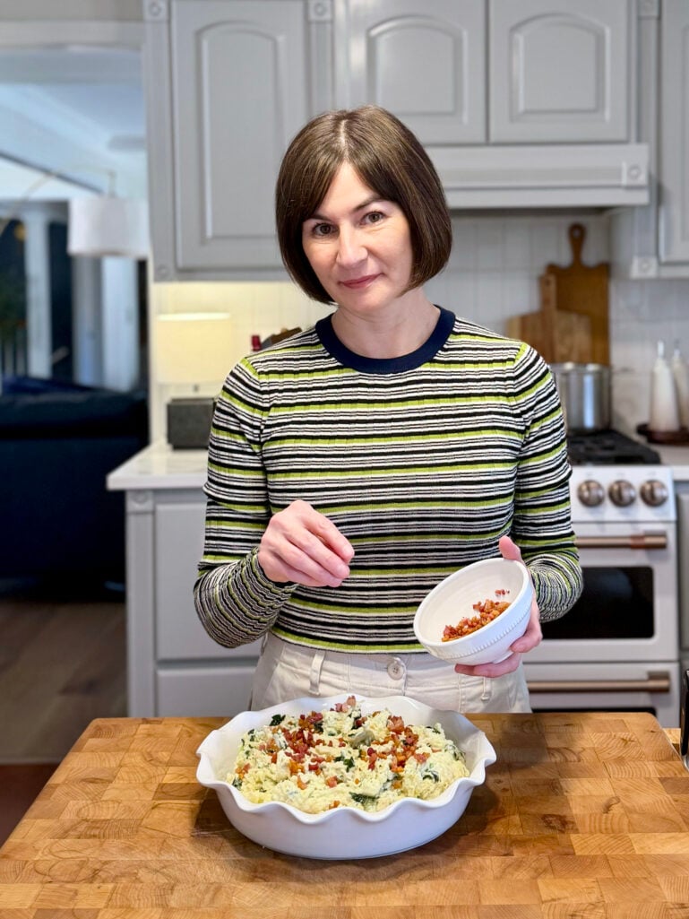 Kelly sprinkling crispy pancetta from a small white bowl over a dish of Colcannon in the kitchen, wearing her green striped crewneck sweater