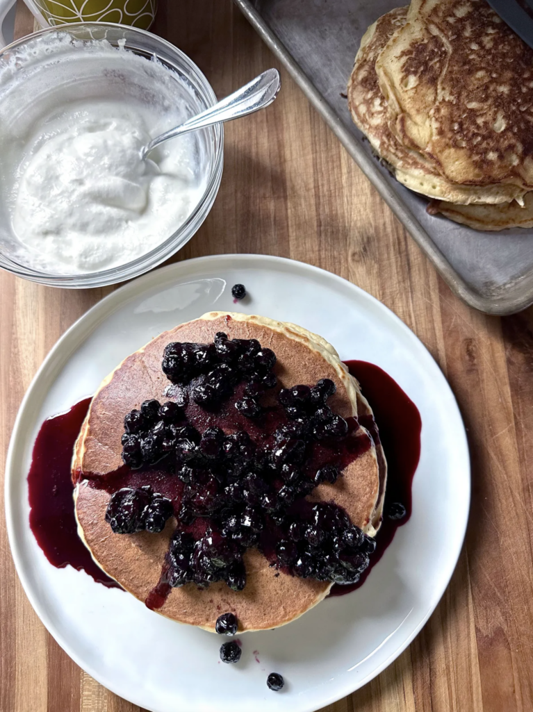 Fluffy lemon ricotta pancakes stacked on a white plate and topped with warm blueberry compote, with extra pancakes on a sheet pan and a bowl of whipped ricotta on a wooden countertop.