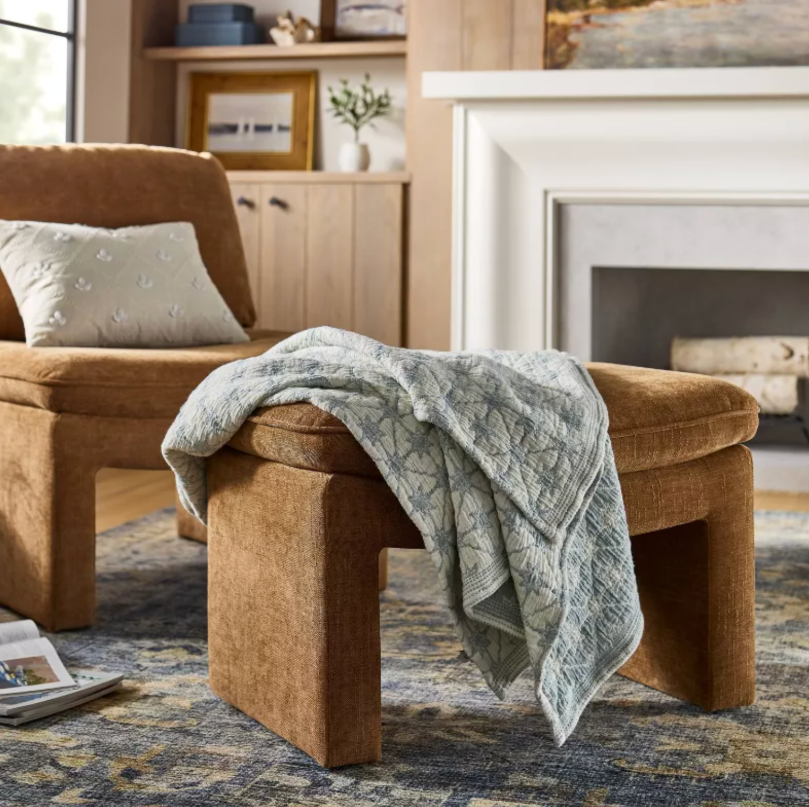 Cozy living room with a warm brown upholstered chair and matching ottoman, draped with a soft textured throw, set against a fireplace and wood shelving with neutral decor.