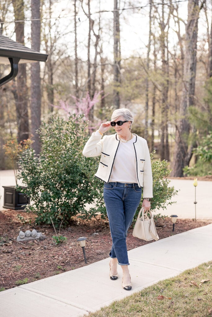 Beth wearing a cream tweed jacket with black trim and gold buttons, white tee, cuffed straight-leg jeans, and beige and black cap-toe block heel slingbacks, carrying a cream structured top-handle bag and wearing a gold chain necklace and black sunglasses