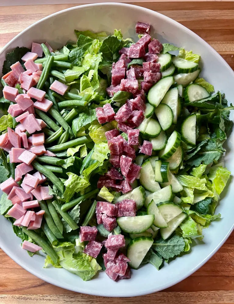 Overhead view of diced ham and salami cubes added in columns over the greens, beans, and cucumbers