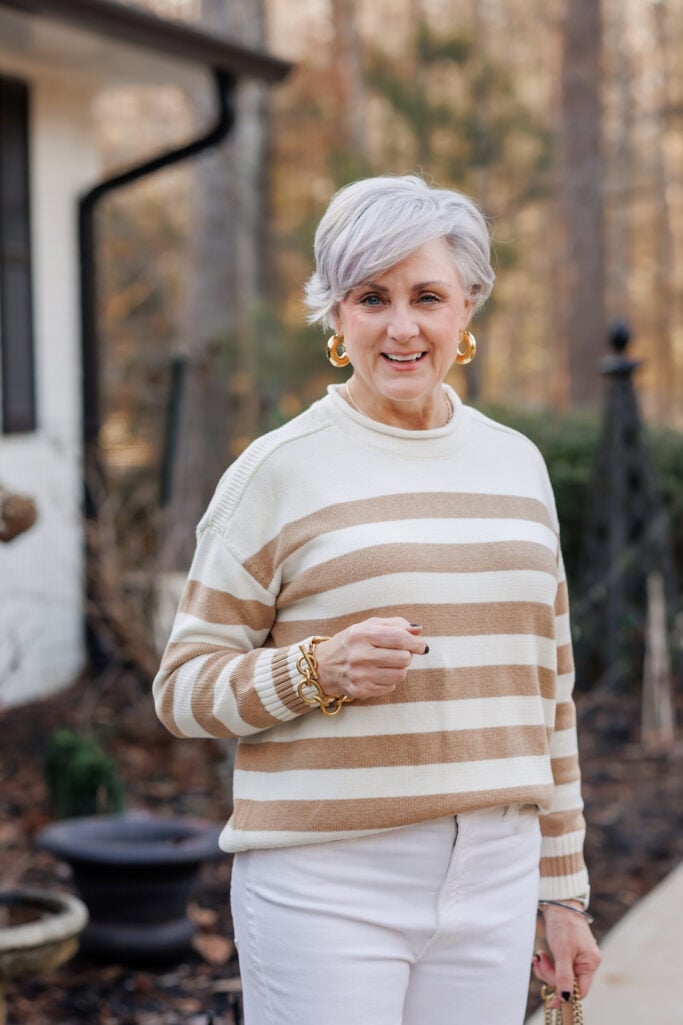 Woman over 50 with silver hair wearing camel and cream striped crewneck sweater with white jeans, chunky gold hoop earrings, gold chain link bracelet