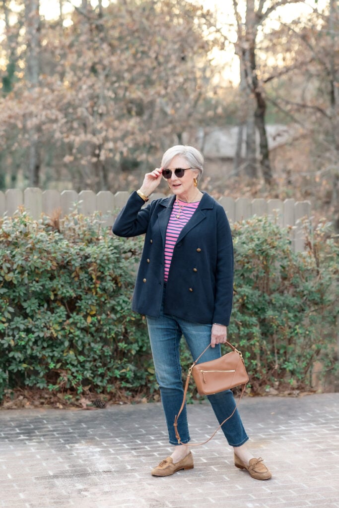 Woman over 50 with silver hair wearing a navy double-breasted sweater blazer over a pink and navy striped long-sleeve tee with slim straight-leg medium-wash jeans, tan suede tassel loafers, cognac patchwork leather crossbody bag, gold hoop earrings, gold bracelet, and tortoiseshell sunglasses, standing on a brick driveway