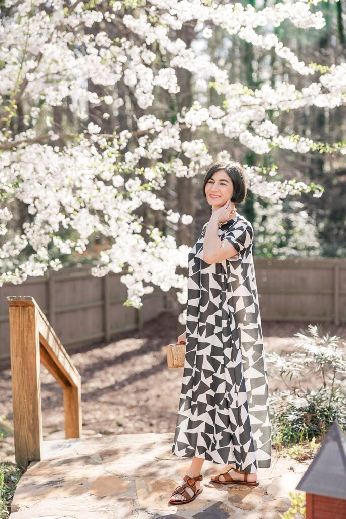 Kelly in Banana Republic Trapeze Maxi Dress black and white geometric print, smiling in sunlit spring garden with white flowering cherry trees in background, gold beaded necklace and brown strappy sandals — petite spring maxi dress