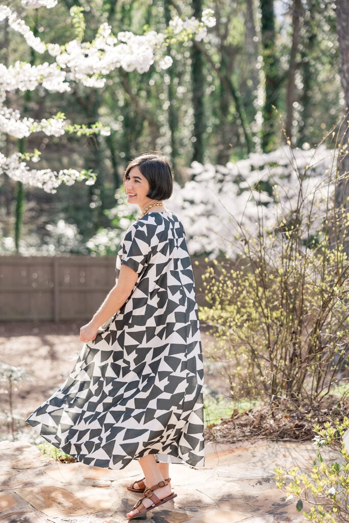 Kelly twirling in Banana Republic Trapeze Maxi Dress showing full black and white geometric skirt in motion, spring cherry blossoms in background, brown strappy leather sandals — petite maxi dress movement