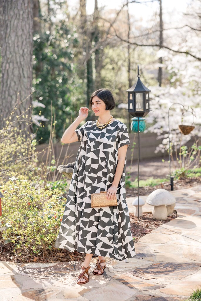 Kelly in Banana Republic Trapeze Maxi Dress geometric print, smiling in spring garden with sunlight filtering through flowering trees, natural woven clutch and brown leather buckle sandals — petite spring outfit