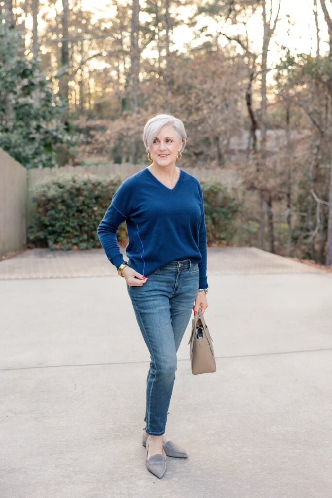 Woman over 50 with silver hair wearing a navy blue V-neck cashmere sweater with slim straight-leg medium-wash jeans, gray suede pointed-toe loafers, taupe leather mini tote bag, gold hoop earrings, and gold bracelet stack, standing on a driveway with trees in the background