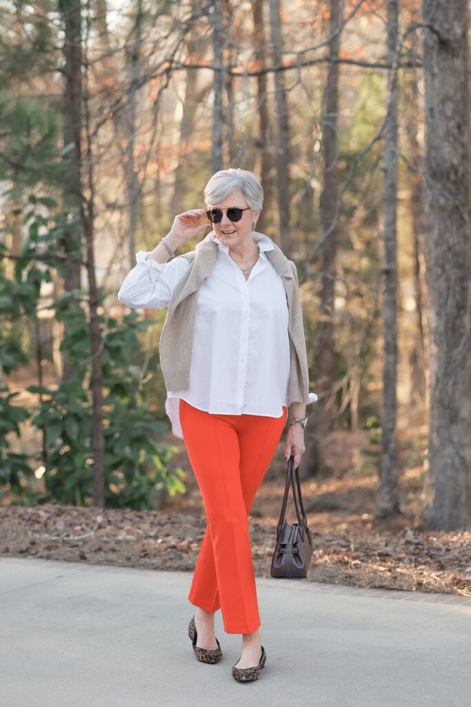 Woman over 50 with silver hair wearing white cotton button-down shirt with oatmeal sweater draped over shoulders, bright orange cropped pants, leopard print ballet flats, brown leather bag, aviator sunglasses