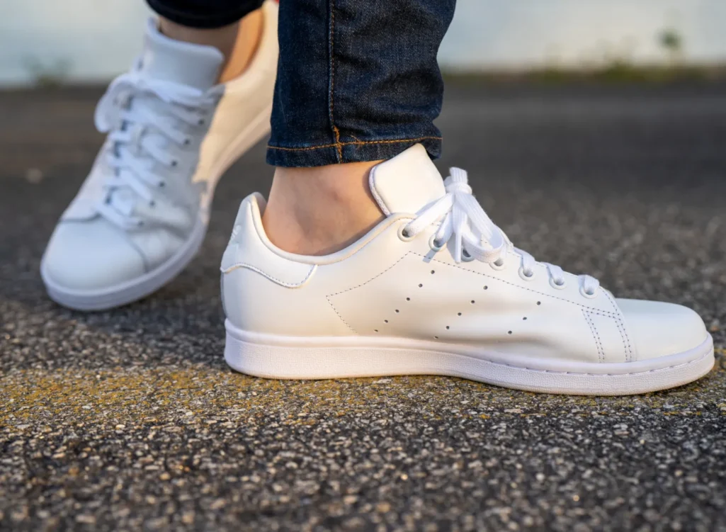 Close-up of woman's feet wearing clean white leather lace-up sneakers with dark wash cuffed jeans on a pavement surface — white leather sneakers spring 2026, best white sneakers for women over 50