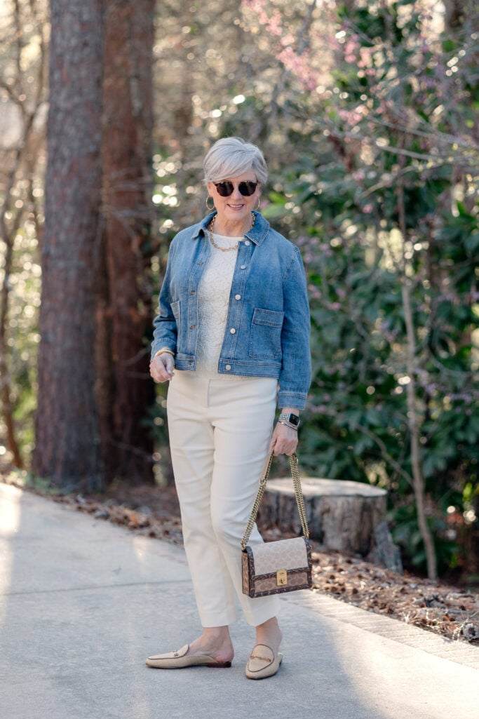 Woman over 50 with silver hair wearing a medium-wash cropped denim jacket over a cream lace tank top with ivory kick flare pants, tan bit loafers, and a neutral Coach canvas chain bag, walking on a tree-lined path in early spring — tonal cream and denim outfit, women over 50