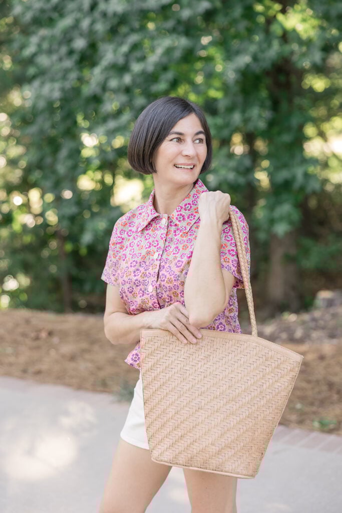 Woman with dark bob haircut wearing a pink and orange floral short-sleeve button-down top with white shorts, carrying a structured natural woven straw bucket tote bag with braided handles — spring bucket bag trend 2026