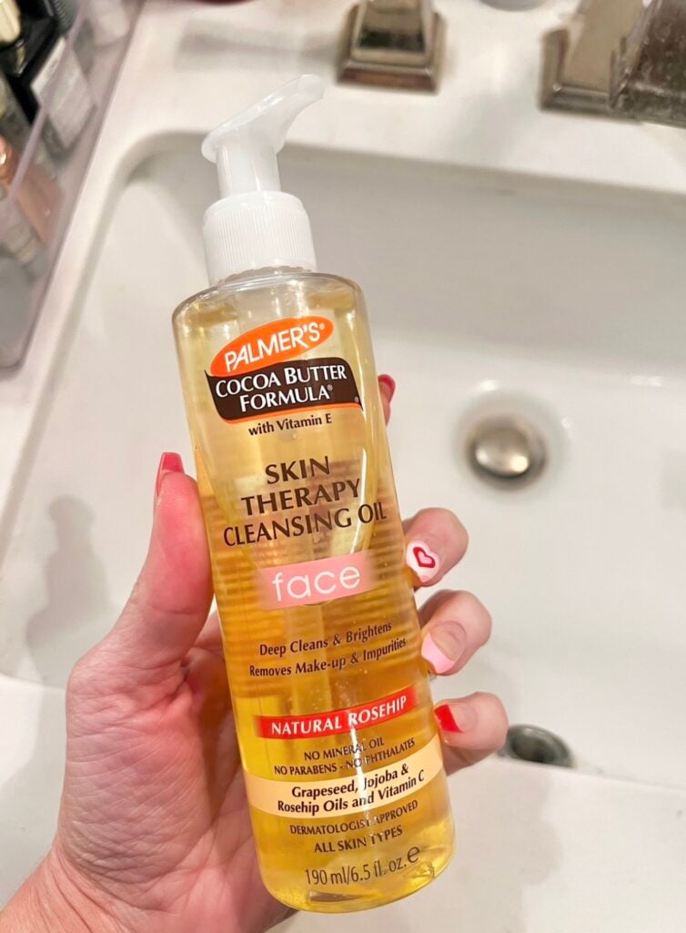 Hand holding clear pump bottle of Palmer's Cocoa Butter Formula Skin Therapy Cleansing Oil for Face showing golden amber oil formula, Natural Rosehip label visible, red and white heart nail art on fingers