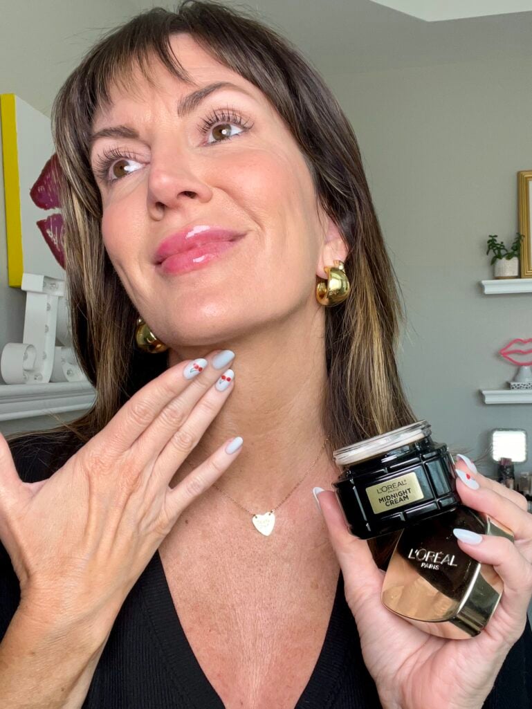 Woman applying L'Oreal Paris Midnight Cream to neck and décolletage while holding open black jar, wearing black V-neck blouse, gold heart pendant necklace and gold bubble earrings, white nails with red cherry nail art, colorful room décor including pink lip wall art in background
