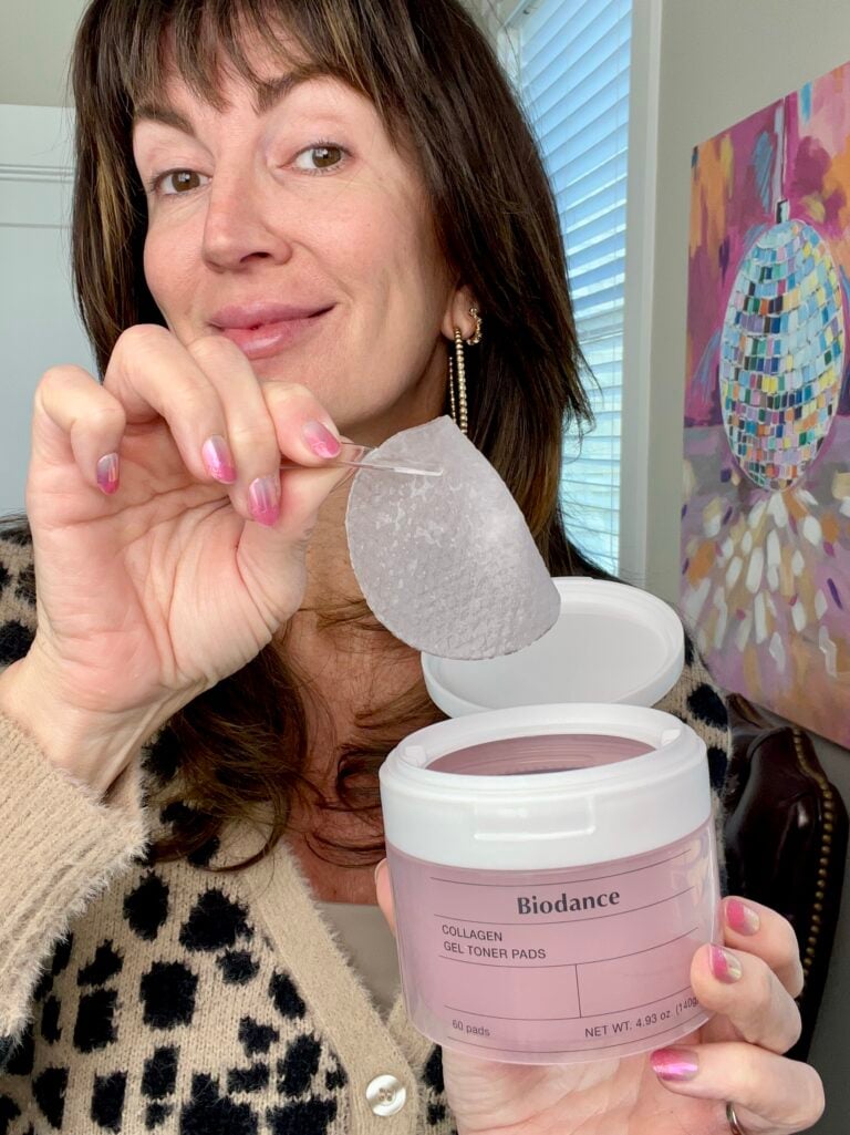 Woman holding open white container of Biodance Collagen Gel Toner Pads and lifting one translucent circular pad between fingers, wearing leopard print cardigan and gold chain hoop earrings, pink ombré nails, colorful disco ball painting in background