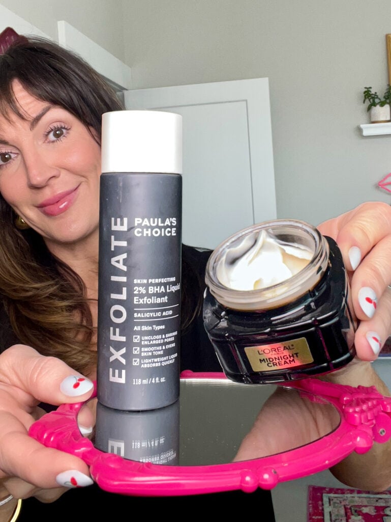 Woman peeking from behind Paula's Choice Skin Perfecting 2% BHA Liquid Exfoliant bottle and open jar of L'Oreal Paris Midnight Cream held in front of a hot pink ornate mirror, white nails with red cherry accent, black top and gold hoop earrings